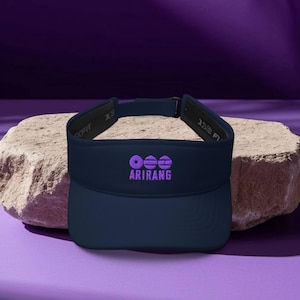 May include: A navy blue visor with the word "ARIRANG" embroidered in purple. The visor has a curved brim and an adjustable strap. The visor is displayed on a light-colored, textured rock against a purple background.