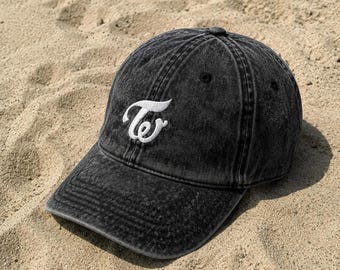 Può includere: Un cappellino da baseball grigio antracite con effetto lavaggio acido e un logo ricamato in bianco sul pannello frontale. Il logo è composto da una lettera T stilizzata e una w minuscola intrecciate. Il cappellino ha una visiera curva.