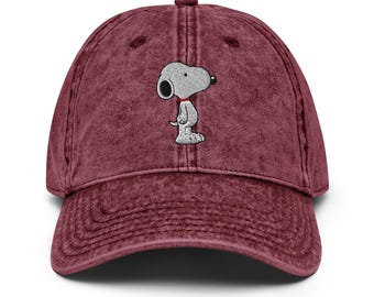Può includere: Un cappellino da baseball color marrone scuro con una texture slavata e vissuta. Il pannello frontale presenta un ricamo di un cane bianco dei cartoni animati con un orecchio nero, naso nero e un sottile collare rosso, visto di profilo.