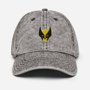 May include: A grey acid-wash baseball cap featuring an embroidered yellow and black graphic of a masked character face on the front panel. The cap has a curved brim, a button on the top, and stitched eyelets for ventilation on the crown panels.
