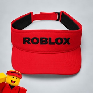 May include: A bright red visor featuring the word ROBLOX embroidered in black capital letters across the front panel. The visor includes a black interior lining and an adjustable strap with a black buckle closure at the back for a secure fit.