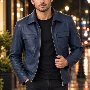 May include: A navy blue leather jacket featuring a classic collar, a full-length silver-tone metal zip, and two chest pockets with flaps. The jacket is worn over a plain black t-shirt and dark-coloured trousers, showcasing a structured design.