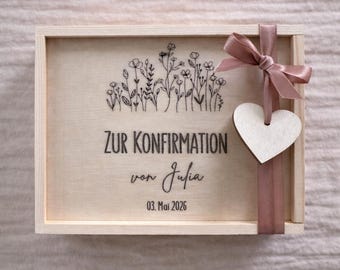 May include: A rectangular light-colored wooden box featuring a black line-art floral illustration and the text "ZUR KONFIRMATION von Julia 03. Mai 2026". A dusty rose ribbon is tied around the right side with a small wooden heart attached.