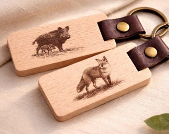 May include: Two wooden keychains with brown leather straps and brass rings. One keychain features a detailed engraving of a fox, and the other depicts a boar with piglets. The keychains are on a cream-colored surface.