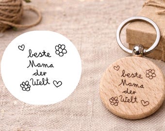 May include: A wooden keychain with the text "beste Mama der Welt" (best mom in the world) engraved on it, along with flower and heart designs. The keychain has a silver ring and is next to a gift box.