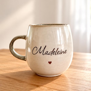 Könnte beinhalten: Ein cremefarbener Keramikbecher mit bauchigem Körper und gesprenkeltem Henkel. Auf der Vorderseite steht der Name Madeleine in dunkelbrauner Schreibschrift, platziert über einem kleinen, ausgefüllten roten Herzsymbol in der Mitte.