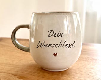 May include: A round, off-white ceramic mug with a curved handle. The front features the black text 'Dein Wunschtext' written in a cursive script, positioned above a small, solid dark red heart icon centered on the lower portion of the mug.