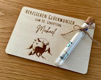 May include: A wooden greeting card with the text "HERZLICHEN GLÜCKWUNSCH ZUM 30. GEBURTSTAG Michael" and a deer silhouette. A glass tube tied with twine contains rolled-up bills, resting on the card. The card and tube are on a wooden surface.