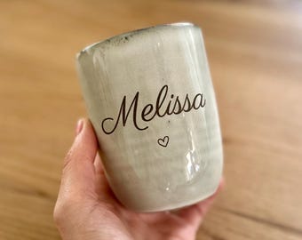 May include: A light gray ceramic mug with a speckled finish and a dark rim. The name Melissa is printed in black cursive script on the front, positioned above a small, simple black heart outline centered on the body of the mug.