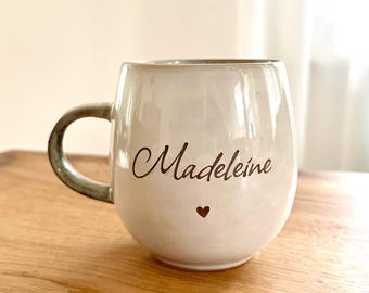 May include: A round, off-white ceramic mug with a glossy finish and a curved handle. The front of the mug features the name "Madeleine" written in a dark brown cursive script, positioned above a small, solid dark brown heart icon.