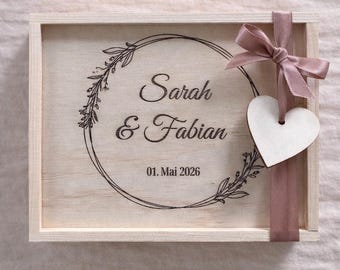 May include: A wooden box with a light brown finish, featuring a circular floral design with the names "Sarah & Fabian" and the date "01. Mai 2026". A pink ribbon and a white heart-shaped tag are attached to the box.