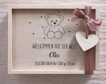 May include: A light wooden box with a teddy bear illustration and the text "WILLKOMMEN AUF DER WELT Cleo 29.12.2025 | 08:34 Uhr | 3267 gr. | 51 cm". A pink ribbon and a wooden heart are attached to the box. The box is a baby keepsake.