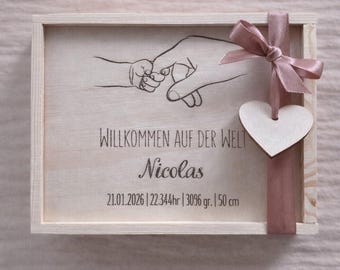 May include: A wooden keepsake box with a hand-drawn illustration of an adult and baby's hands holding each other. The box is tied with a pink ribbon and a wooden heart. Text on the box reads "WILLKOMMEN AUF DER WELT Nicolas 21.01.2026 | 22.344hr | 3096 gr. | 50 cm."