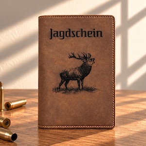 Könnte beinhalten: Ein brauner Dokumentenhalter aus Leder mit dem Wort Jagdschein und einer Illustration eines Hirsches. Der Einband hat sichtbare Nähte an den Rändern. Mehrere messingfarbene Metallhülsen sind links neben dem Halter positioniert.