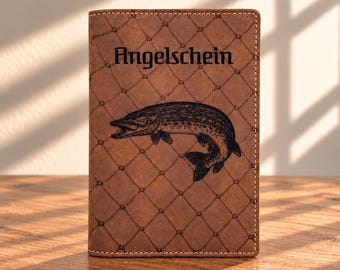 May include: A brown leather wallet featuring a diamond-shaped grid pattern and a black engraved illustration of a pike fish. The word "Angelschein" is printed in a bold, black gothic-style font above the fish illustration on the front cover.
