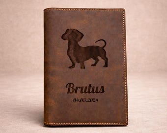 May include: A brown leather passport holder featuring a dark engraved silhouette of a dachshund dog. Below the dog, the name Brutus is engraved in a script font, followed by the date 04.03.2024. The cover has visible stitching along the edges.