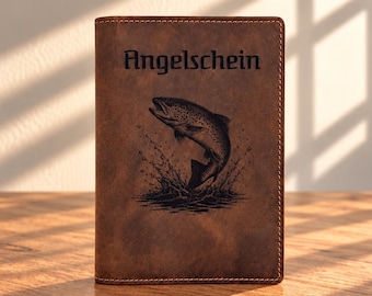 May include: A brown leather document holder featuring a dark engraved illustration of a fish jumping out of water. The word "Angelschein" is embossed in a black gothic-style font above the fish graphic. The cover has visible stitching along the edges.