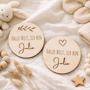 Könnte beinhalten: Zwei runde Holzschilder zur Babyankündigung. Eines zeigt ein Zweig-Design, das andere ein Herz. Beide haben den Text "Hallo Welt, Ich bin Julia" und den Namen "Julia" in einer Schreibschrift. Ein Holzstern und andere Babyartikel sind im Hintergrund.