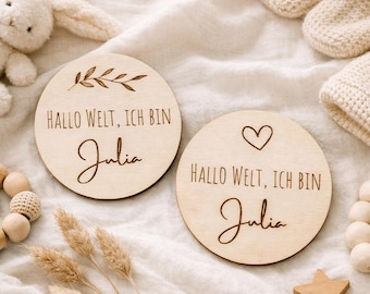 May include: Two round wooden baby announcement plaques. One features a sprig design, the other a heart. Both have the text "Hallo Welt, Ich bin Julia" and the name "Julia" in a script font. A wooden star and other baby items are in the background.