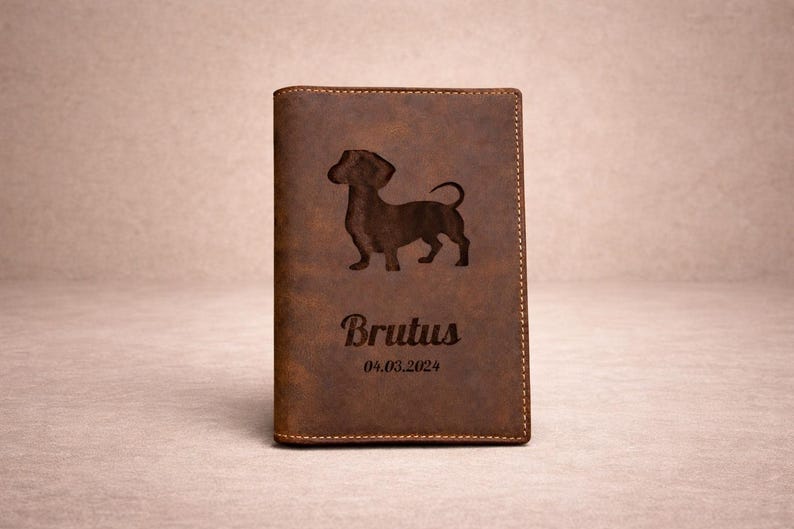 Peut inclure: Un porte-passeport en cuir marron avec la silhouette grav&eacute;e d'un chien teckel. Sous le chien, le nom Brutus est grav&eacute; dans une police cursive, suivi de la date 04.03.2024. La couverture pr&eacute;sente des coutures visibles sur les bords.