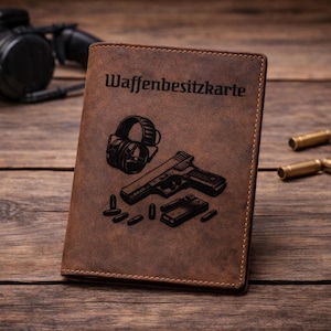 Könnte beinhalten: Ein brauner Leder-Dokumentenhalter mit der Gravur Waffenbesitzkarte, einer Handfeuerwaffe, einem Magazin, mehreren Patronen und einem Gehörschutz. Der Einband weist sichtbare Nähte an den Rändern sowie eine matte, strukturierte Oberfläche auf.