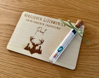 May include: A wooden card with the text "HERZLICHEN GLÜCKWUNSCH ZUR BESTANDENEN JAGDPRÜFUNG! Paul" and a deer silhouette. A small glass vial with a cork stopper and green ribbon is next to the card.