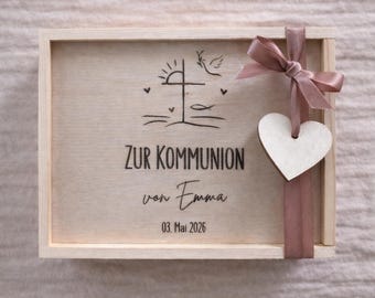 May include: Wooden box with a light brown finish, featuring a cross, dove, and sun design. The box is tied with a mauve ribbon and a wooden heart. Text on the box reads "ZUR KOMMUNION von Emma 03. Mai 2026".