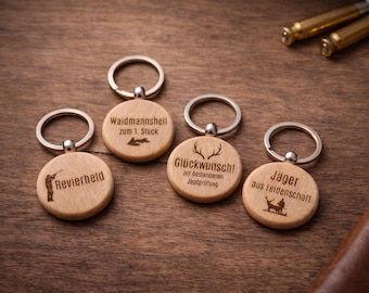 May include: Four wooden keychains with silver rings, each engraved with German text and hunting-themed illustrations. The keychains are round and made of light-colored wood, resting on a dark wooden surface.