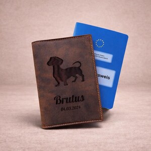 Peut inclure: Une couverture de passeport en cuir marron avec la silhouette grav&eacute;e au laser d'un teckel, le nom Brutus et la date 04.03.2024. Un passeport allemand bleu est partiellement visible derri&egrave;re, montrant l'embl&egrave;me et le texte de l'Union europ&eacute;enne.