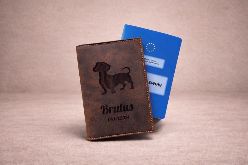 Peut inclure: Une couverture de passeport en cuir marron avec la silhouette grav&eacute;e au laser d'un teckel, le nom Brutus et la date 04.03.2024. Un passeport allemand bleu est partiellement visible derri&egrave;re, montrant l'embl&egrave;me et le texte de l'Union europ&eacute;enne.