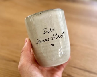 May include: A light gray ceramic mug with a speckled finish and a dark rim. The front features the black printed text "Dein Wunschtext" in a cursive font, positioned above a small, solid black heart icon centered on the mug.