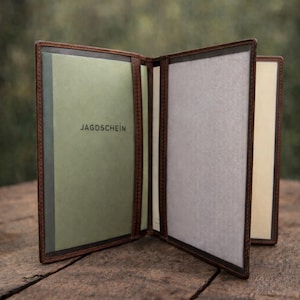 May include: A brown leather document holder with multiple interior sleeves. The left panel features a light green insert with the text JAGDSCHEIN printed in black. The center and right panels contain light gray and cream-colored paper inserts.