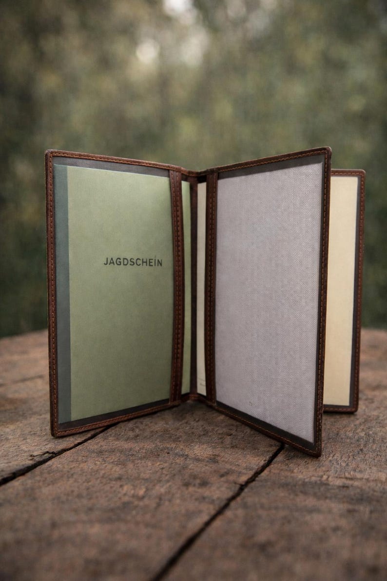 May include: A brown leather document holder with multiple interior sleeves. The left panel features a light green insert with the text JAGDSCHEIN printed in black. The center and right panels contain light gray and cream-colored paper inserts.
