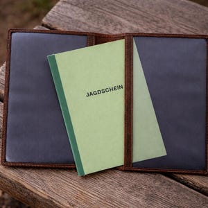 May include: A brown leather document holder with dark interior pockets, containing a light green booklet with the word JAGDSCHEIN printed in black on the front cover. The holder is open, displaying two clear plastic sleeves for document storage.