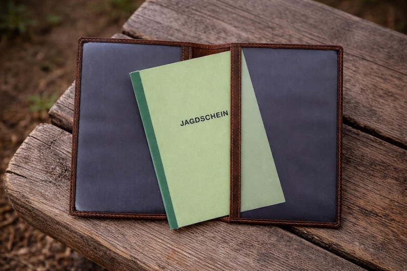May include: A brown leather document holder with dark interior pockets, containing a light green booklet with the word JAGDSCHEIN printed in black on the front cover. The holder is open, displaying two clear plastic sleeves for document storage.