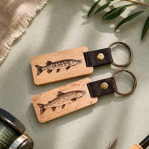 Peut inclure: Deux porte-clés en bois avec des motifs de poissons gravés. Chaque porte-clés comprend une base rectangulaire en bois, une lanière en cuir marron foncé et un anneau métallique. Un porte-clés a un motif de brochet, l'autre de truite. Accessoires sur le thème de la pêche.