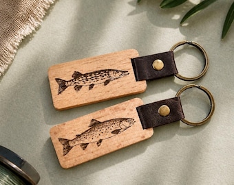 May include: Two wooden keychains with engraved fish designs. Each keychain features a rectangular wooden base, a dark brown leather strap, and a metal ring. One keychain has a pike design, the other a trout. Fishing-themed accessories.