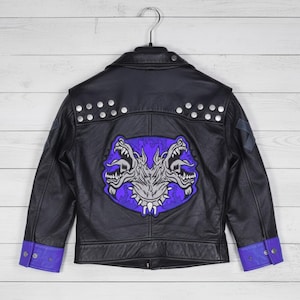 May include: Black leather jacket with silver studs and purple accents. Features a large patch on the back with a snarling two-headed dog design in gray and purple. The jacket is hanging on a metal hanger.