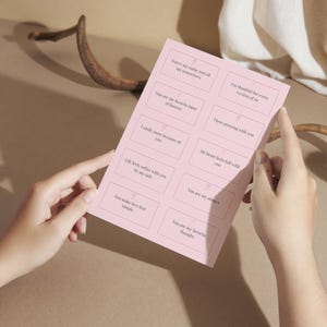 May include: A pink card with various love quotes in a grid layout. The card is held by two hands against a neutral background. Quotes include "You're my today and all my tomorrows" and "You are my favorite kind of forever."