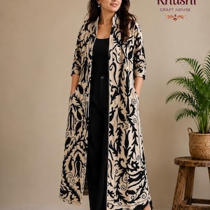 May include: A long, beige open-front coat featuring a bold black floral pattern, worn over a black top and black trousers. The garment has three-quarter length sleeves and side pockets. The text Khushi Craft House is visible in the upper right.