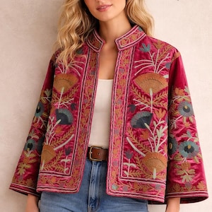 Può includere: Giacca in velluto magenta con ricami floreali. La giacca ha un collo alto e maniche a tre quarti. Il ricamo comprende sfumature di oro, verde e blu. Il marchio "Khushi Craft House" è visibile in alto a destra.