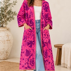Könnte beinhalten: Ein langer, vorne offener Mantel in leuchtendem Pink mit dunkelvioletten Blumenstickereien und einem weiß-rosa gestreiften Saum. Das Kleidungsstück hat zwei große aufgesetzte Vordertaschen und wird über einem weißen Oberteil und hellblauen Jeans getragen.