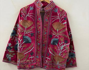 May include: A magenta velvet jacket with floral embroidery in shades of brown, blue, and green. The jacket has a mandarin collar and is open in the front, hanging on a wooden hanger. The brand logo "Khushi Craft House" is visible in the upper right corner.