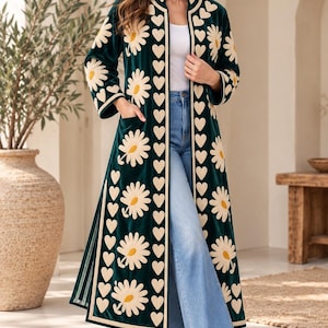 Pode incluir: Um casaco comprido tipo duster em veludo verde-escuro com bordados florais de margaridas em tom creme e uma borda vertical com padrão de corações ao longo da abertura frontal. A peça inclui mangas compridas, bolsos laterais e gola alta.