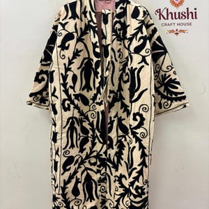 May include: A long, open-front robe in cream fabric featuring bold black floral and vine embroidery. The garment has wide sleeves and a red and white striped lining visible at the collar. The text Khushi Craft House is printed in the top right corner.