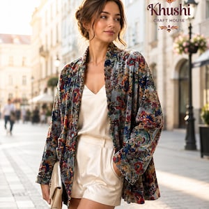 May include: A woman in a floral print jacket, white top, and shorts, holding a beige clutch. The jacket has a colourful floral pattern on a grey background. The image is taken outdoors on a city street.