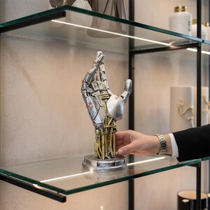 Könnte beinhalten: Eine silber-goldene Roboterhand-Skulptur mit komplizierten mechanischen Details. Die Skulptur steht auf einem Glasregal und zeigt ihre metallische Oberfläche und ihr futuristisches Design. Die Hand ist in einer Geste gehalten, die eine Begrüßung oder ein Angebot andeutet.