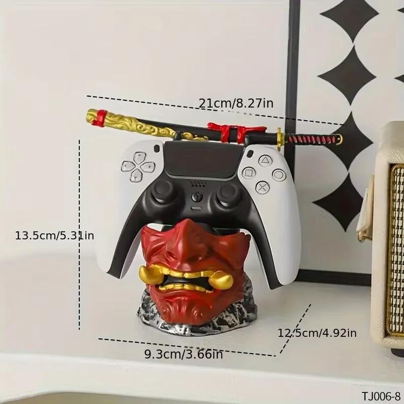 K&ouml;nnte beinhalten: Ein wei&szlig;-schwarzer Videospiel-Controller auf einem roten Oni-Masken-St&auml;nder. Ein Miniatur-Katana-Schwert, 21 cm lang, liegt auf dem Controller. Der St&auml;nder ist 9,3 cm breit und 12,5 cm hoch.