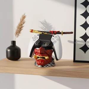 Könnte beinhalten: Ein weiß-schwarzer Videospiel-Controller liegt auf einem rot-goldenen Oni-Maskenständer. Ein dekoratives Katana liegt auf dem Controller. Der Ständer steht auf einem Holzregal mit einer schwarzen Vase und einem gerahmten Kunstdruck im Hintergrund.