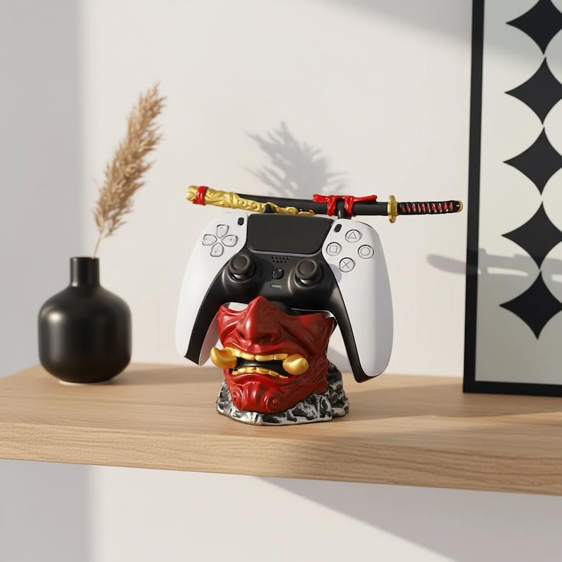 K&ouml;nnte beinhalten: Ein wei&szlig;-schwarzer Videospiel-Controller liegt auf einem rot-goldenen Oni-Maskenst&auml;nder. Ein dekoratives Katana liegt auf dem Controller. Der St&auml;nder steht auf einem Holzregal mit einer schwarzen Vase und einem gerahmten Kunstdruck im Hintergrund.